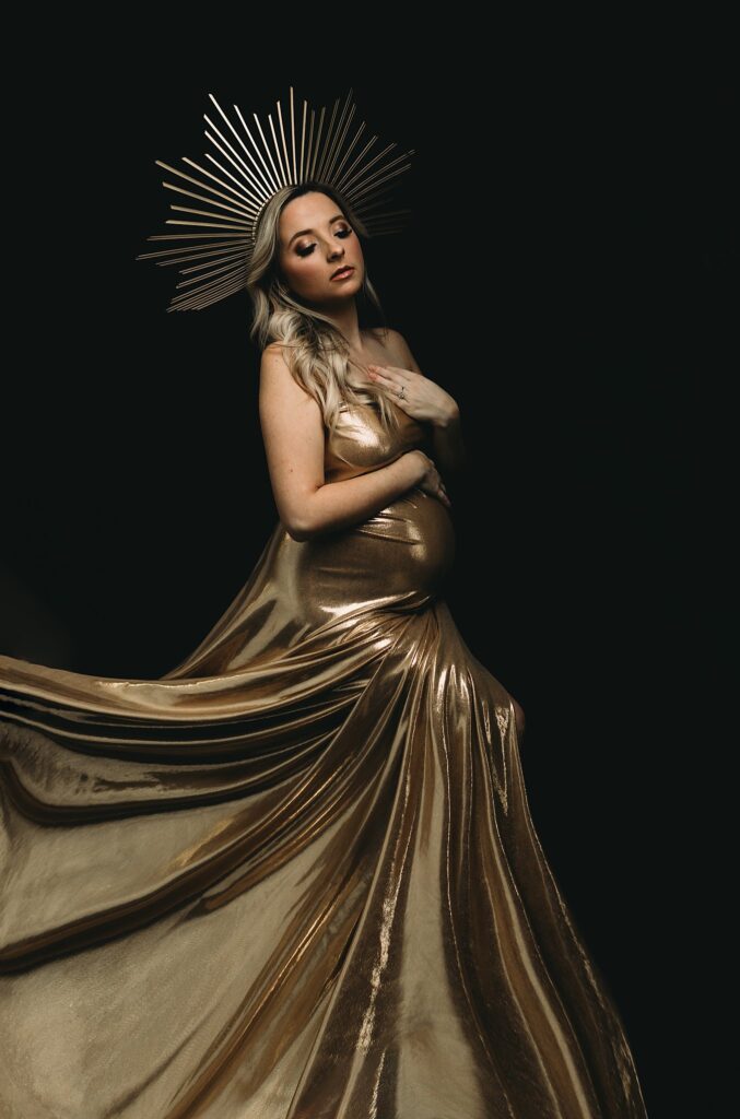 Pregnant woman dressed as a golden goddess with a gold gown and crown against a black background for her maternity photoshoot