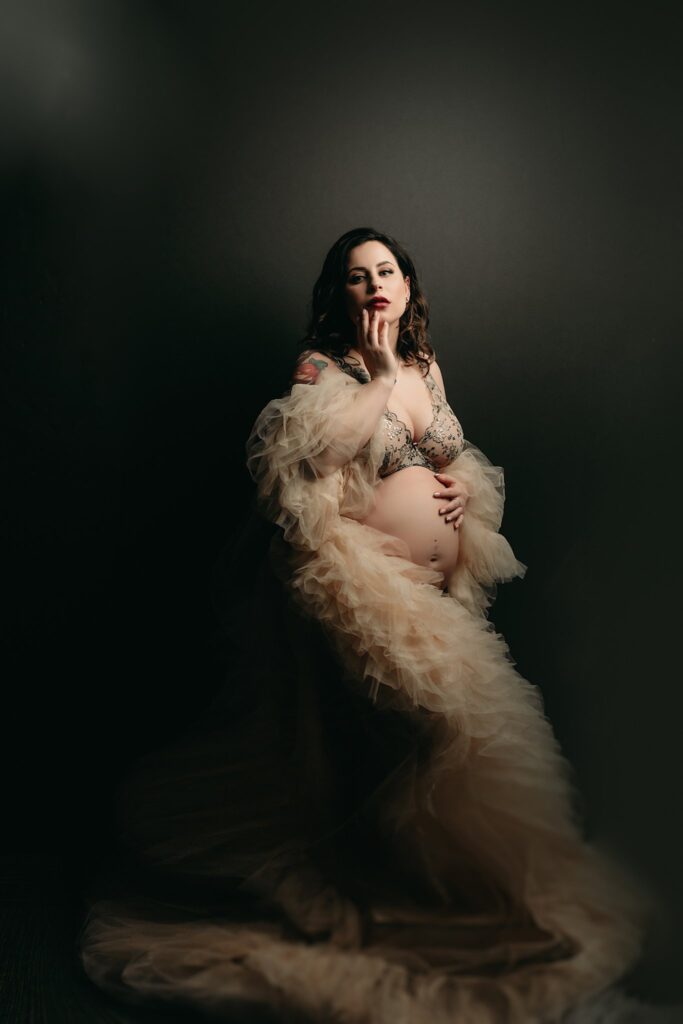 Expecting mother in a ruffled, sheet negligee and bralette against dark background in natural lighting for a romantic feel