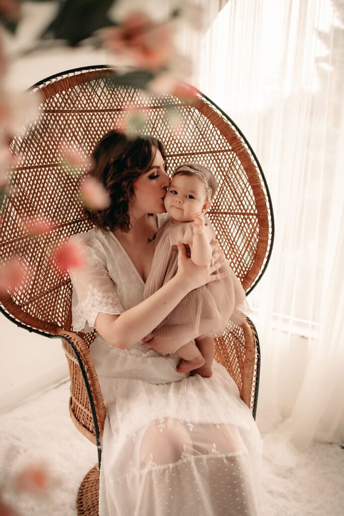 A mother and with infant daughter sitting in a wicker chair, the child embraced by its mother in a room with natural light and florals