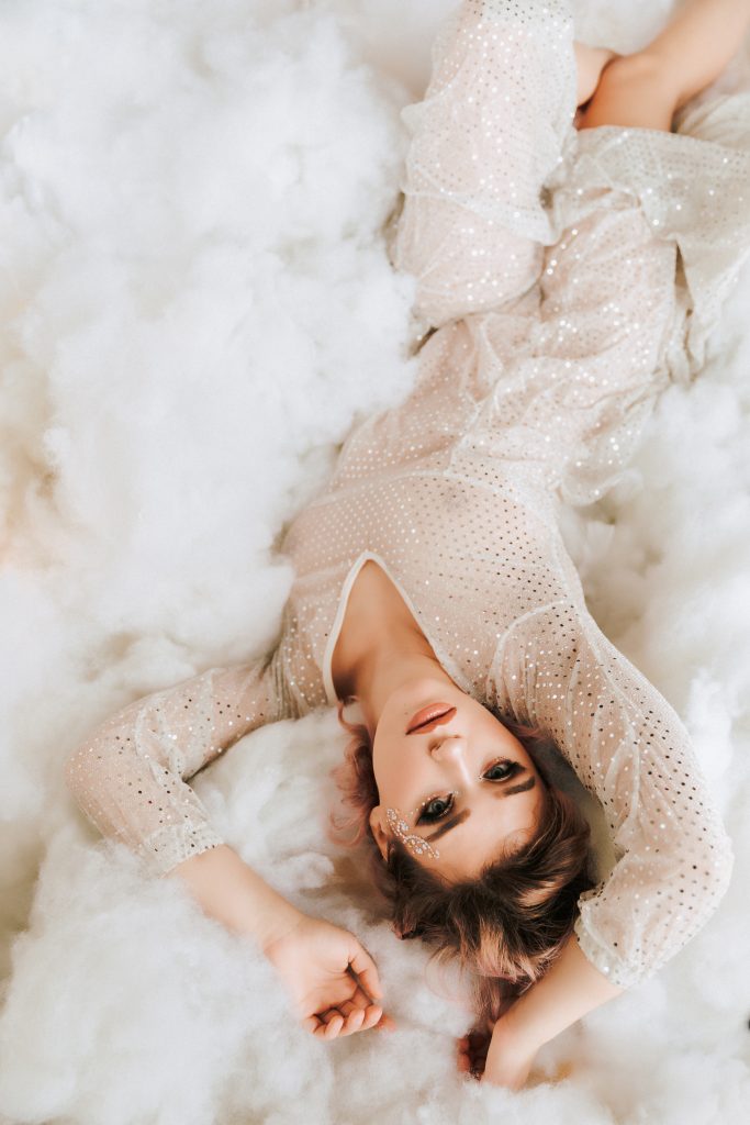 beautiful woman in see through glittery dress and glitter on face staring into camera with pink lipstick wearing glittery dress laying in airy light and bright fluffy cloud scene upside down Dijana Szewczyk photography Portland Oregon Boudoir Washington
