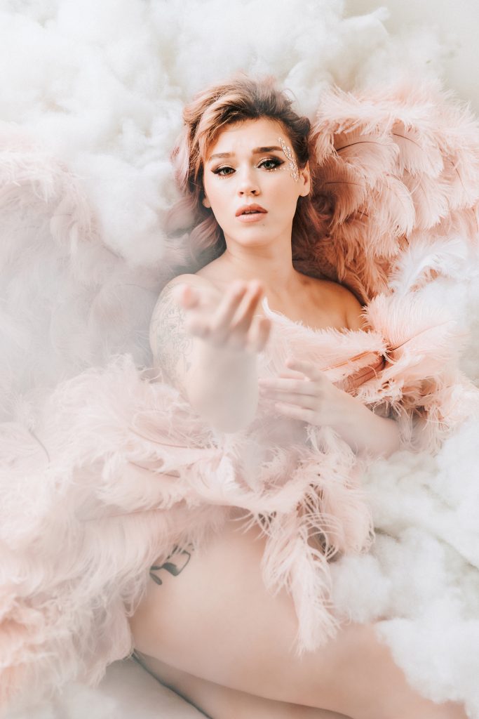beautiful woman laying in light bright and airy fluffy cloud background in pink angel wings with tattoos and glitter on face looking into camera with hand extended Dijana Szewczyk photography Portland Oregon Boudoir Washington
