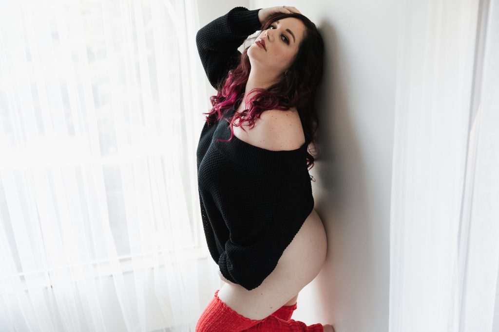white woman with brown hair and peachy lipstick and. pink hair tips looking into camera with hand in hair wearing black sweater and red thigh high socks against white wall in light and airy setting Dijana Szewczyk photography Portland Oregon Boudoir Washington