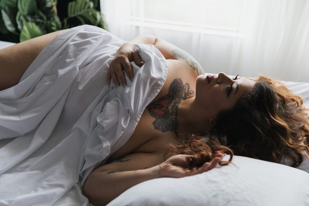 beautiful Latina woman rolling around in white sheets in a light and airy setting sheets draped over body looking into. air red lipstick and tattoos Dijana Szewczyk photography Portland Oregon Boudoir Washington