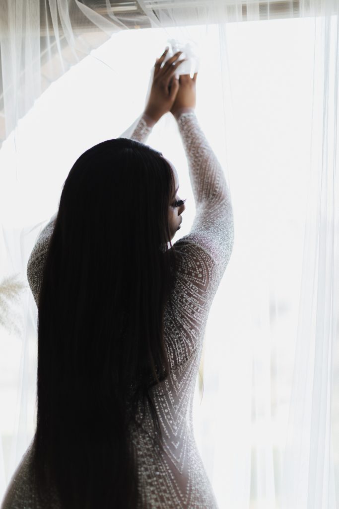 Beautiful African American woman posing in front of lighted and airy window with hands raised and eyes closed looking to side in see through, glittery dress Dijana Szewczyk photography Portland Oregon Boudoir
