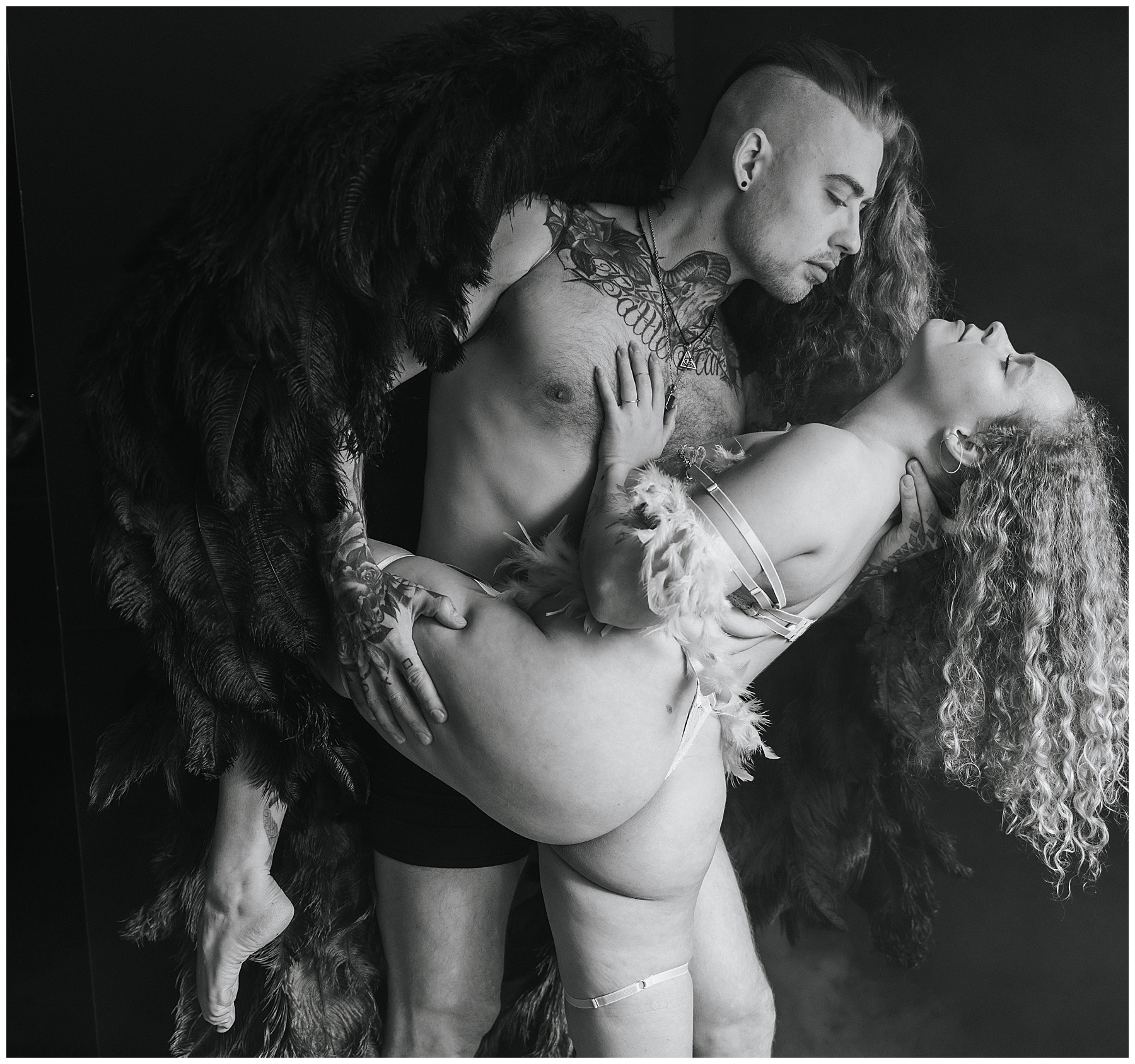 black and white photo, man with partially shaved head and partially long curly hair wearing black wings and black briefs with tattoos covering right hand and arm and across top of chest and neck is holding a woman against his body, his head is bent forward, woman is wearing white thong garment with straps draped down her arm, left leg bent and raised being held by man's right arm, her left arm is bent next to her body and placed on man's chest, his left arm is holding her neck as she tips her head backwards