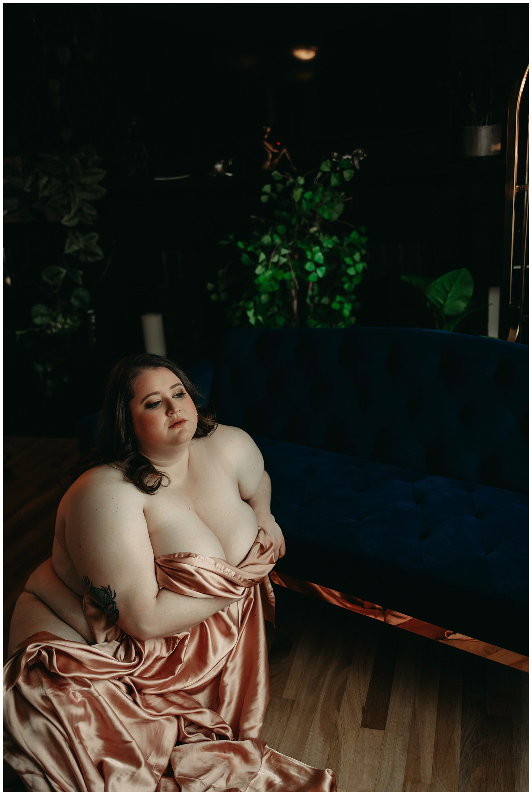 a plus sized white woman is sitting on the floor naked with a silky blanket covering the front half of her body while learning on a blue couch, there are green plants in the background. she has brown hair and has a serious face.
