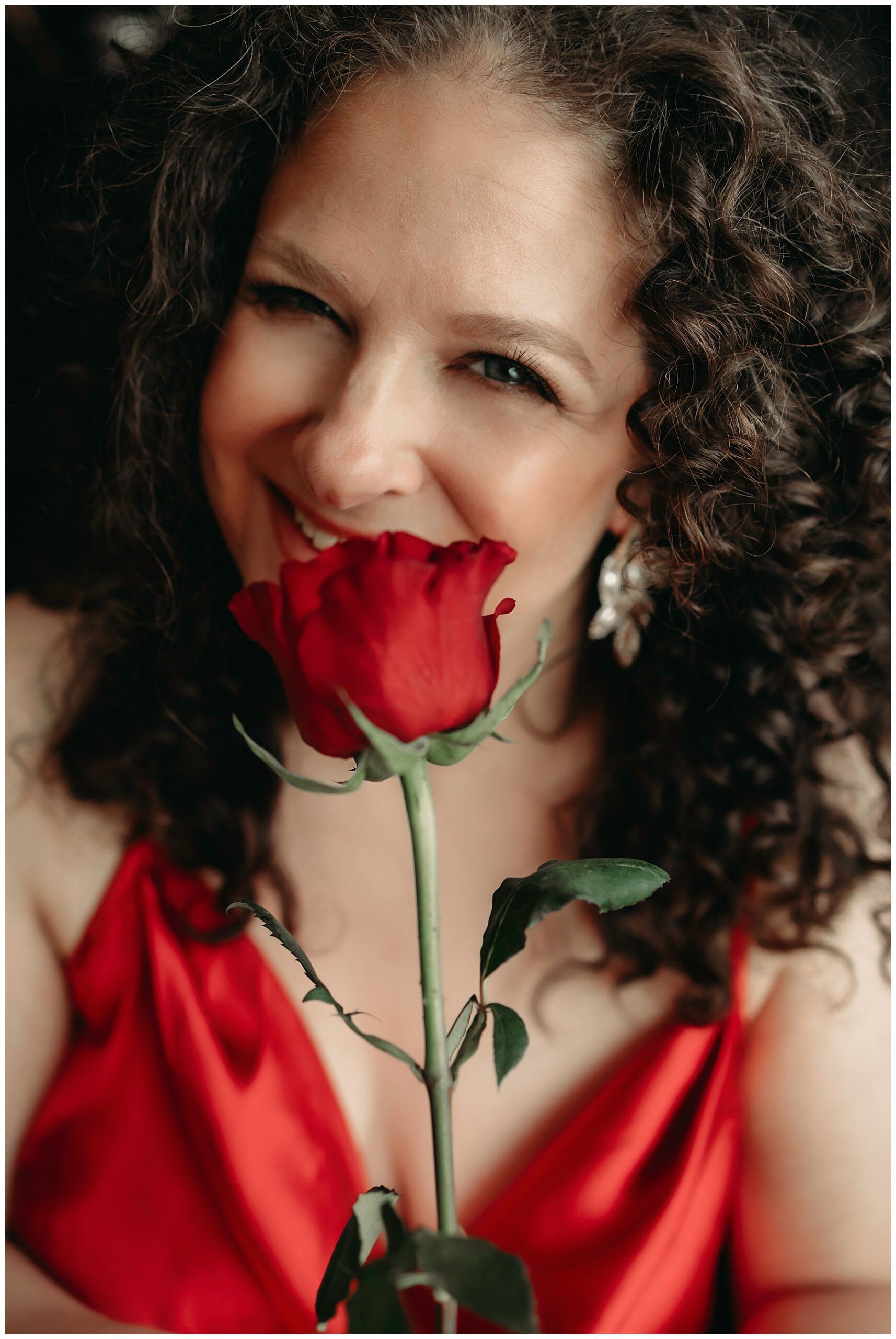 a close up of a woman with brown curly hair in a red dress holding a red rose looking at the camera and smiling, she is happy